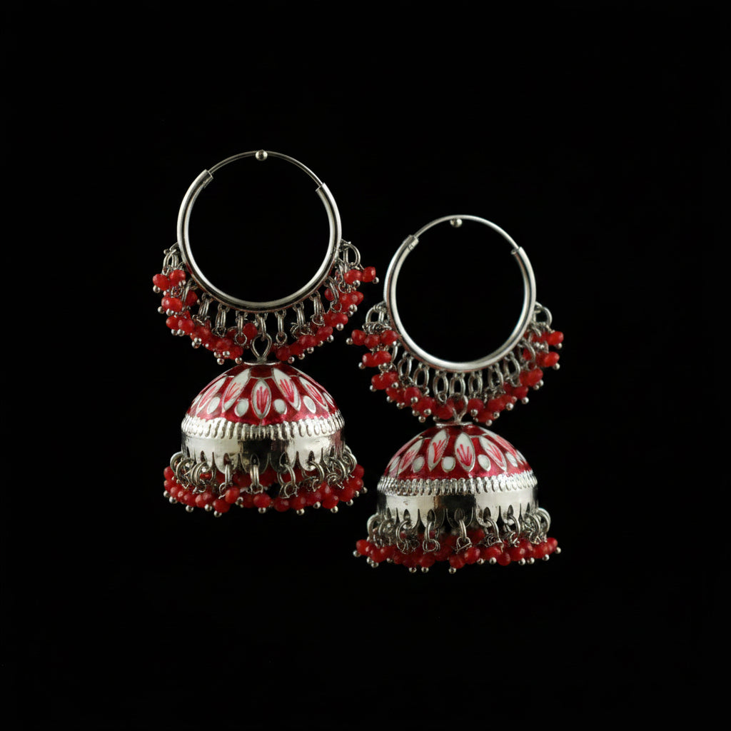 A pair of silver-toned Indian jhumka earrings with red bead fringe and red-and-white patterned bell-shaped drops, displayed on a black stand against a gray background.