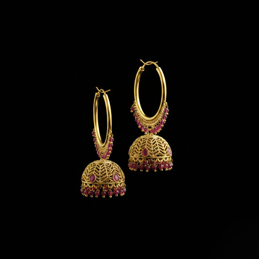A pair of gold-toned traditional hoop earrings with dangling filigree jhumka-style bells and small pink gemstone accents, displayed on a black jewelry stand against a dark gray background.