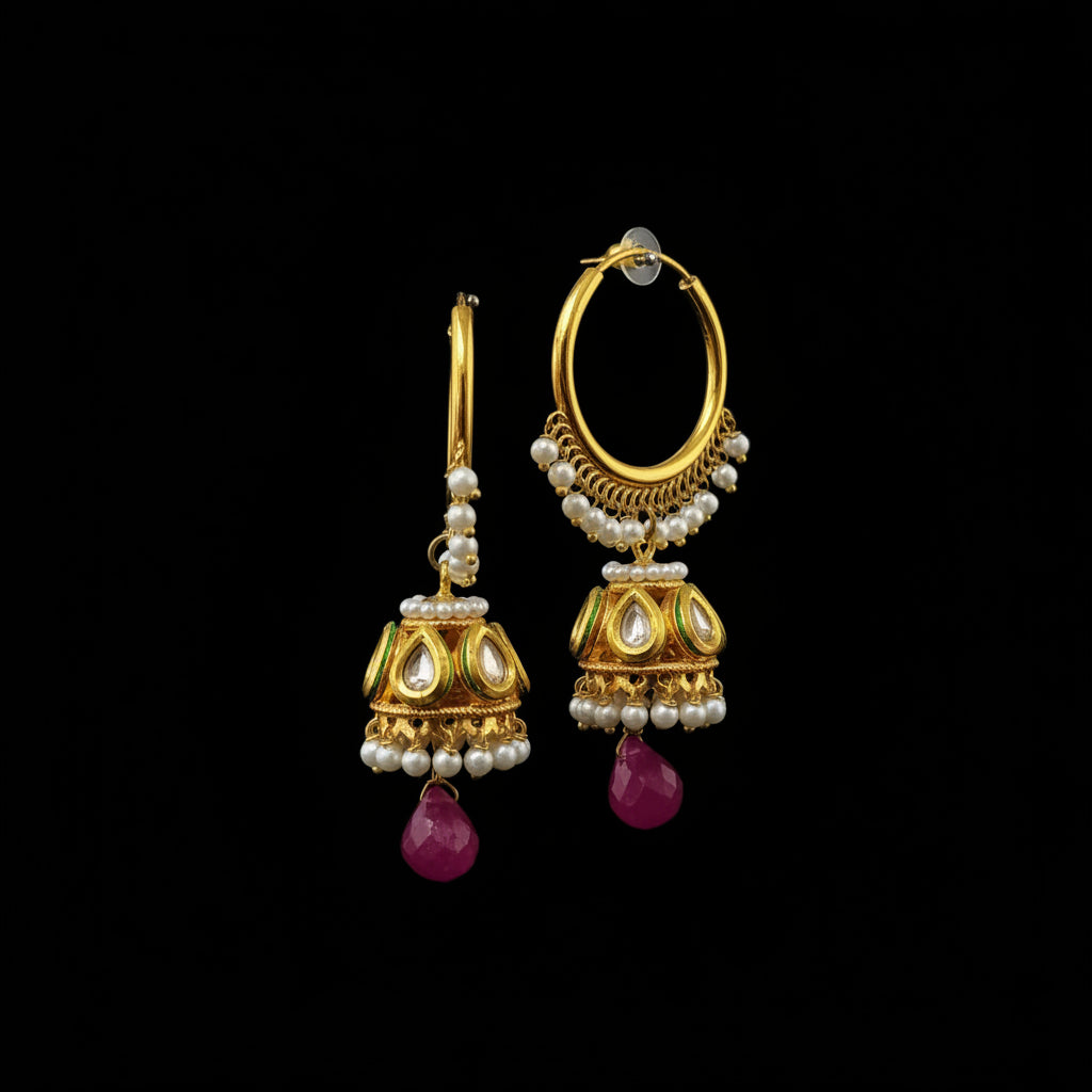 A pair of gold-toned Indian jhumka earrings with white pearl bead borders, gold-toned kundan motifs, and dangling magenta pink teardrop crystals, displayed on a gray stand.