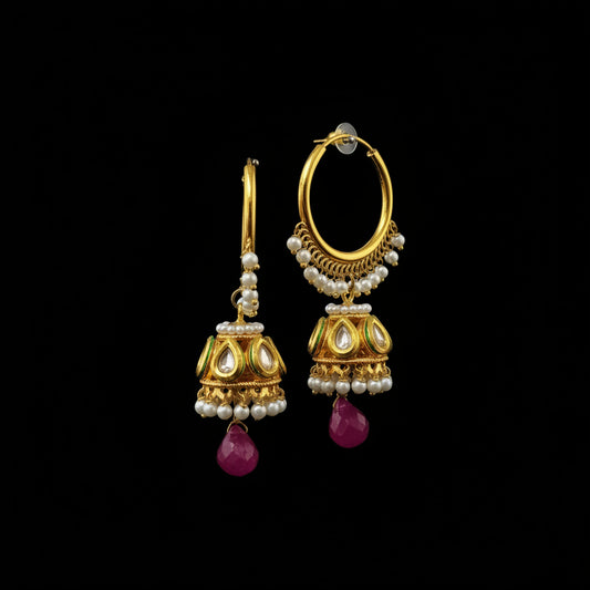 A pair of gold-toned Indian jhumka earrings with white pearl bead borders, gold-toned kundan motifs, and dangling magenta pink teardrop crystals, displayed on a gray stand.