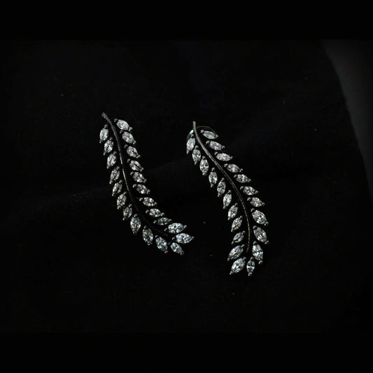 A pair of leaf-shaped drop earrings with marquise-cut clear stones set in a dark metal, displayed on a black velvet surface with the SAHELI MY BEST FRIEND logo in the top right.