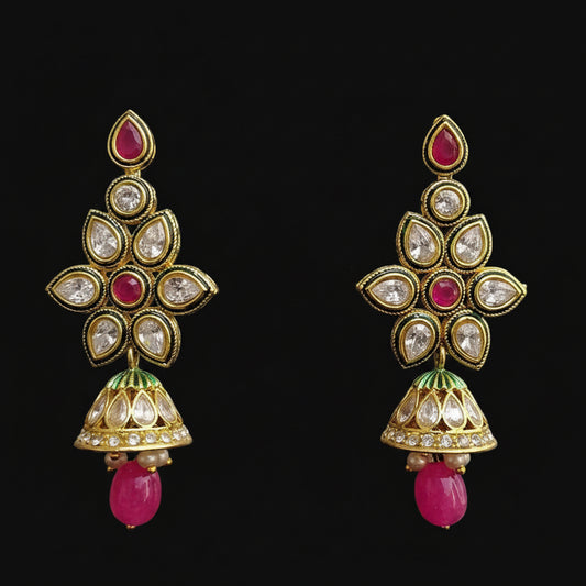 A pair of gold-plated Indian jhumka earrings with pink stone accents, clear crystals, and green enamel details, displayed on a black Y-shaped stand against a gray background. The brand logo 'SAHELI MY BEST FRIEND' is visible at the top.