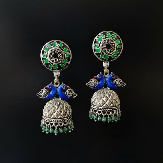 Ornate earrings with blue birds and green accents on a gray stand against a black background.