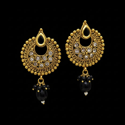 A pair of gold-toned traditional Indian jhumka earrings with black bead details and clear stones, displayed on a black Y-shaped stand against a gray background with the brand logo 'SAHeli My Best Friend' at the top.
