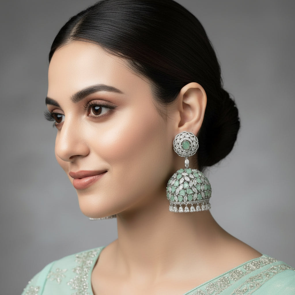 Pair of earrings with green gemstones and silver accents on a black stand, branded 'Saheli'.