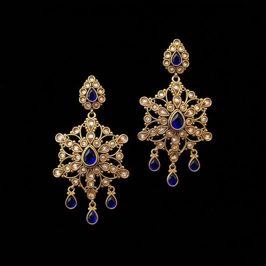 A pair of ornate gold-toned chandelier earrings with deep blue teardrop stones and clear stone accents, displayed on a gray stand against a gray background with the brand name SAHELI MY BEST FRIEND visible at the top.