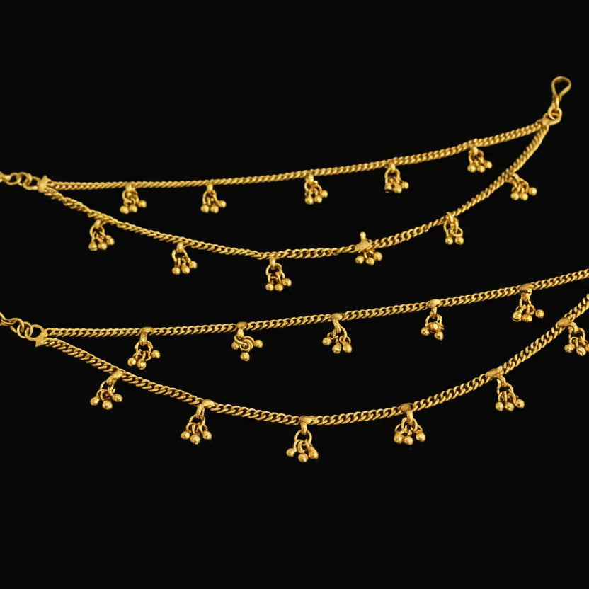 Two gold anklets with dangling charms on a dark background
