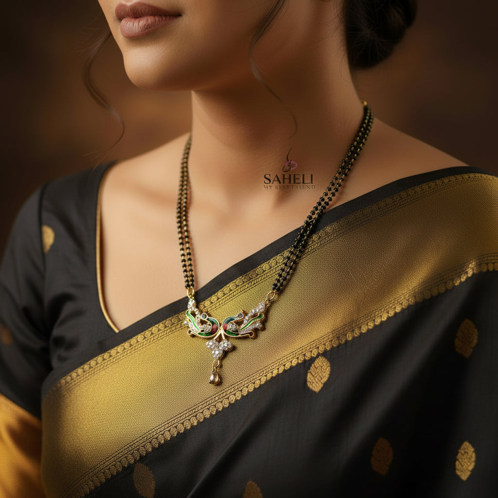 A black beaded necklace with a decorative pendant featuring gold, green, and red enamel details, displayed on a dark reflective surface. The brand logo 'SAHELI' is visible in the upper right.