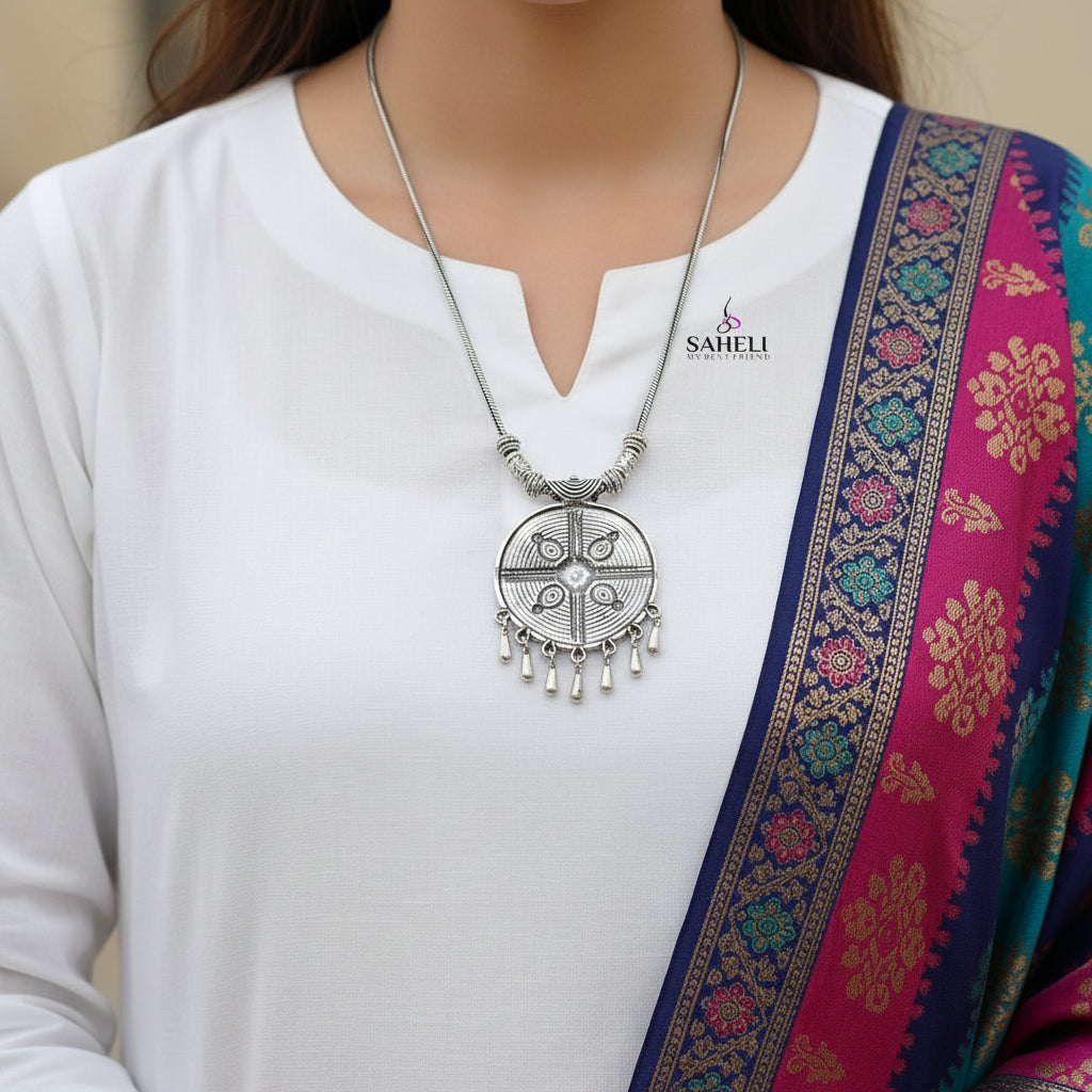 Silver necklace with a decorative pendant on a gray background, branded 'SAHELI'.