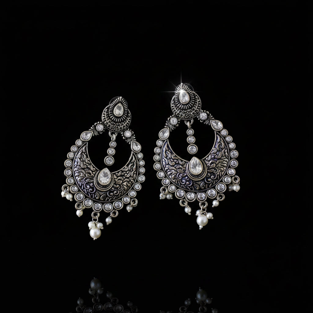 A pair of ornate silver-toned chandelier earrings with intricate filigree detailing, clear rhinestones, and white pearl bead drops, displayed on a dark reflective surface with the SAHELI brand logo above.