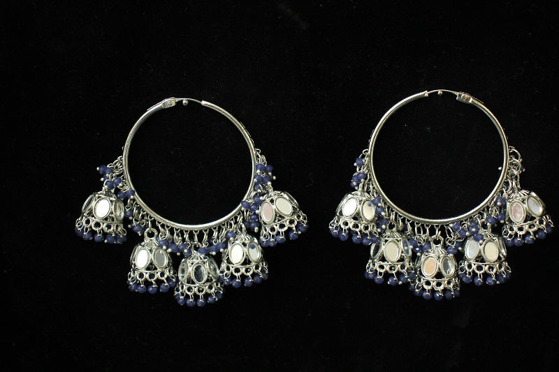 A pair of silver-toned huggie hoop earrings decorated with dangling silver filigree, white stone accents, and small dark blue beads, displayed on a black background.