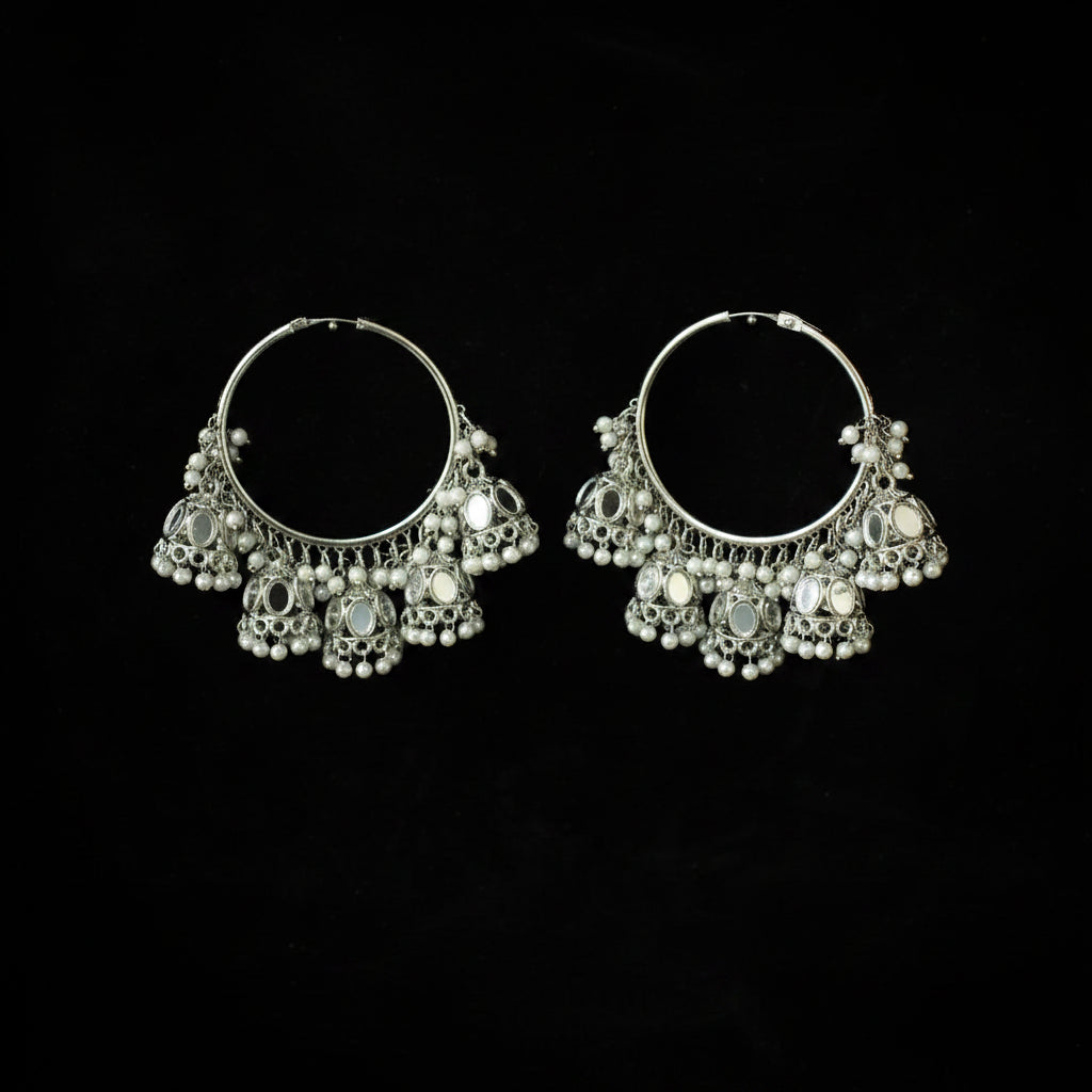 A pair of silver-toned traditional hoop earrings adorned with dangling mirror work, white beads, and small metallic charms, displayed against a solid black background.