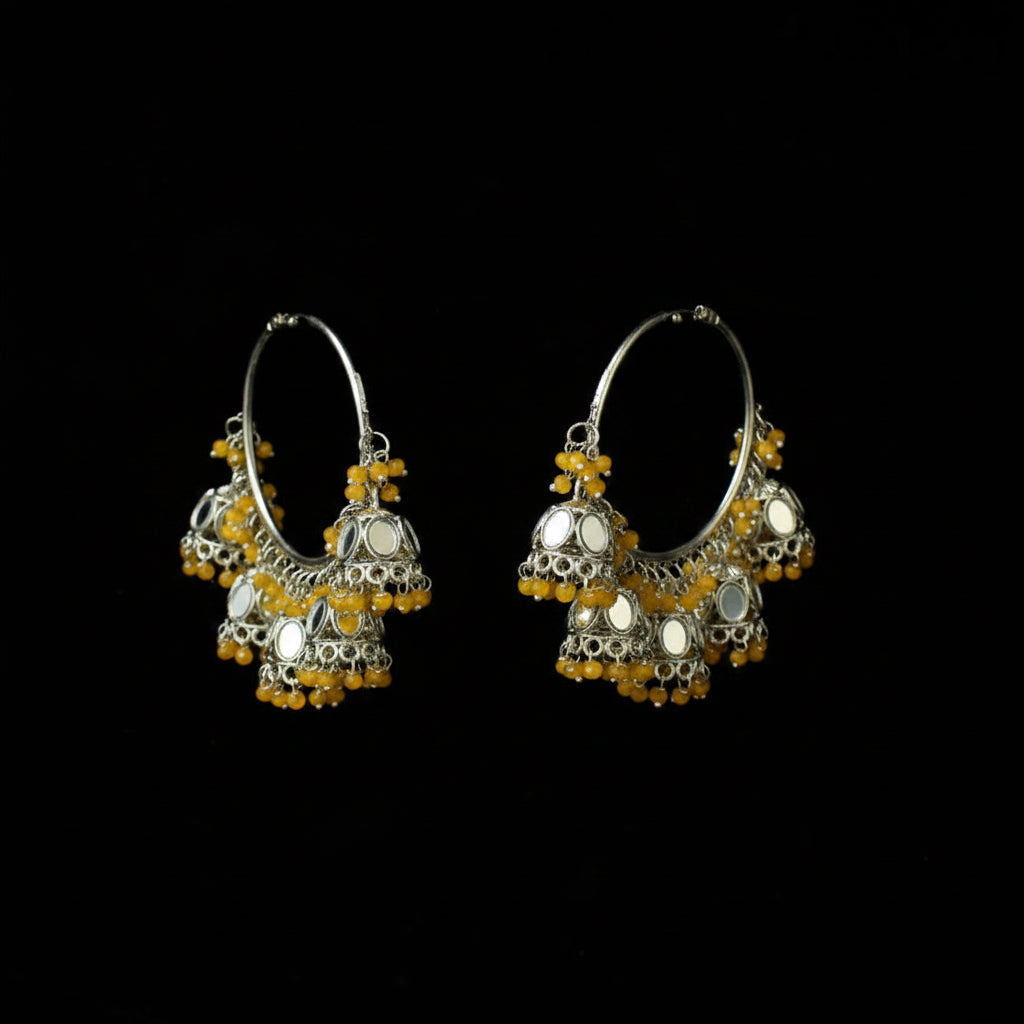 A pair of silver-toned traditional hoop earrings with dangling bell-shaped charms, yellow bead accents, and circular mirror inlays, displayed against a solid black background.