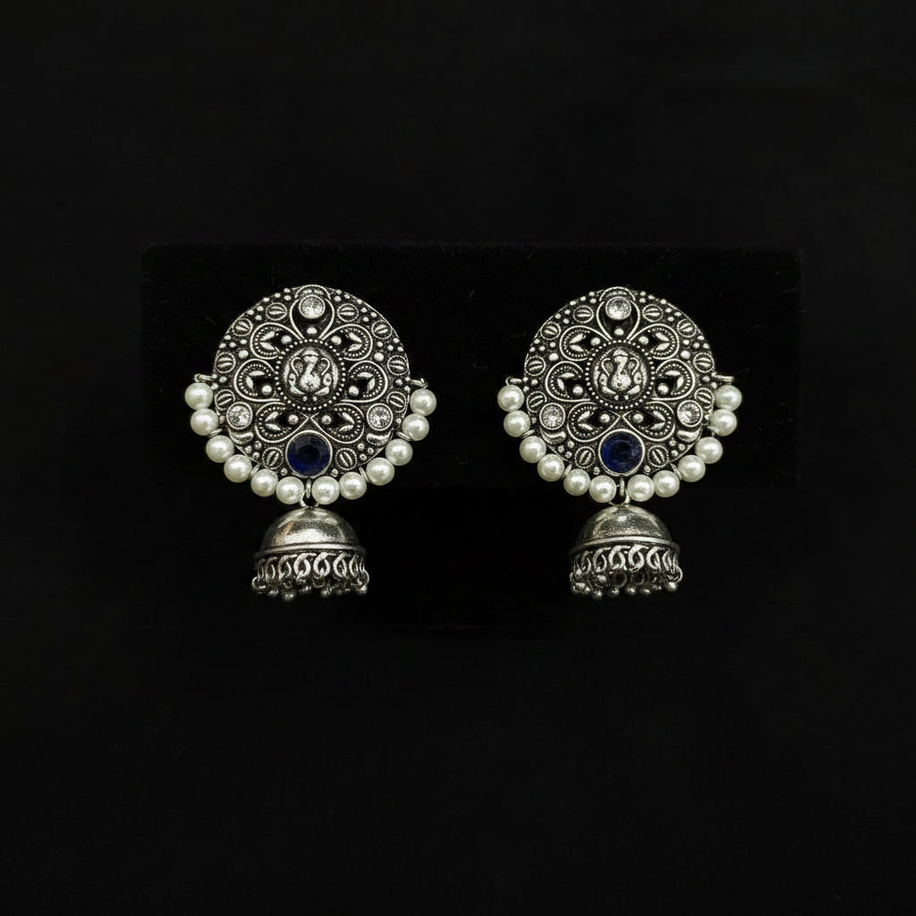 A pair of traditional Indian silver-toned jhumka earrings with intricate filigree work, white faux pearl borders, and dark blue stone accents, displayed on a black textured background.