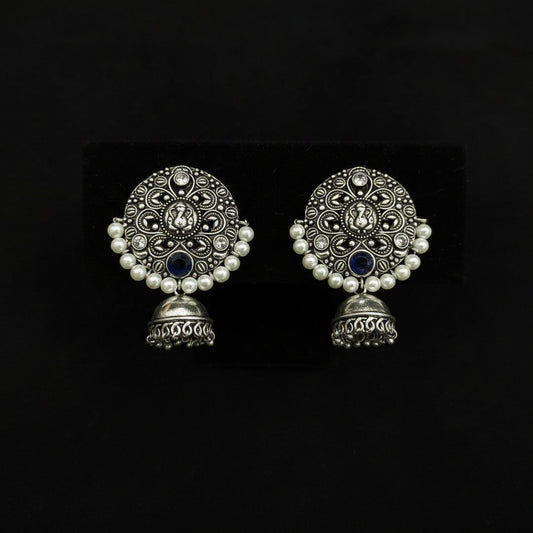 A pair of traditional Indian silver-toned jhumka earrings with intricate filigree work, white faux pearl borders, and dark blue stone accents, displayed on a black textured background.