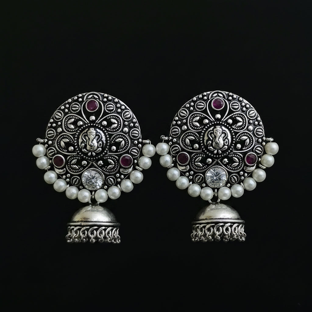 A pair of ornate silver-toned Indian jhumka earrings with intricate filigree work, white pearl bead borders, and small purple gemstone accents, displayed on a black stand against a dark background.