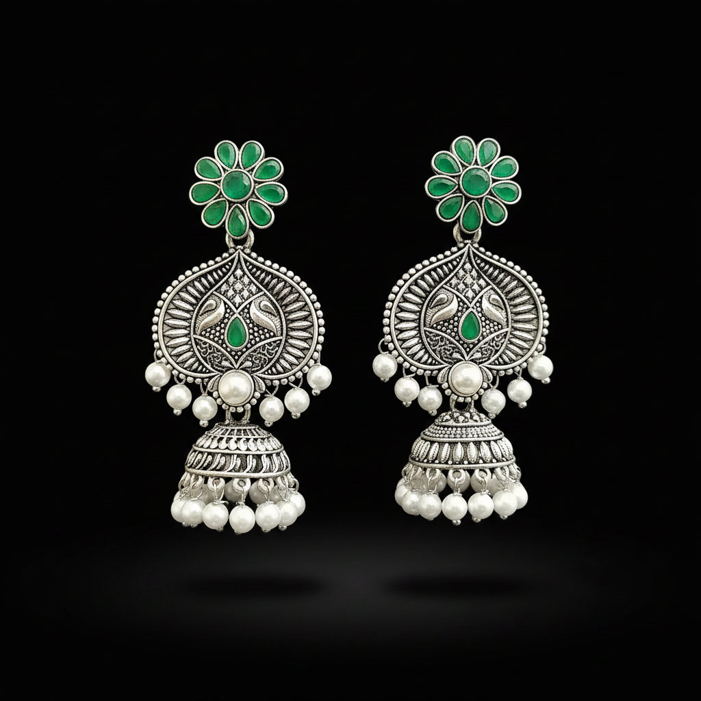 Ornate silver earrings with green gemstones and pearl-like beads on a gray stand.