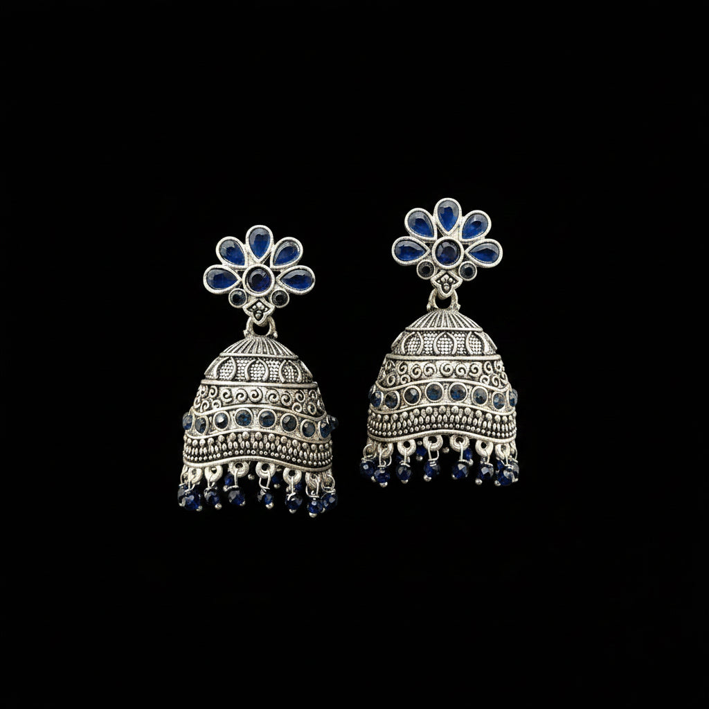 A pair of silver-toned Indian jhumka earrings with blue stone accents, displayed on a gray velvet jewelry stand against a light gray background.