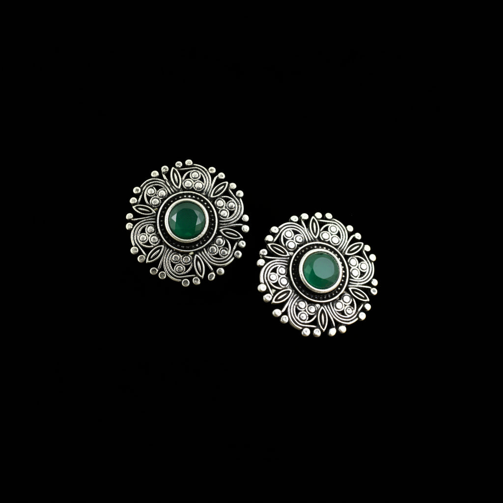A pair of silver-toned filigree stud earrings with oval green stone centers, displayed on a dark reflective surface.