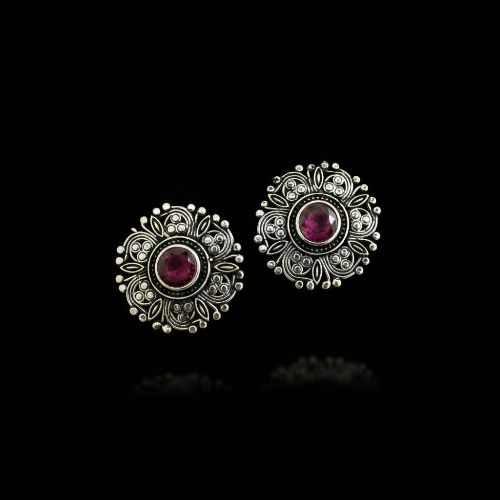 A pair of ornate silver-toned filigree earrings with round red gemstone centers, displayed on a dark reflective surface.