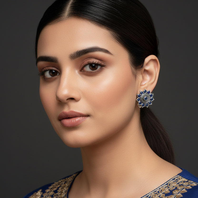 A pair of silver-toned floral stud earrings with deep blue circular stones, displayed on a dark reflective surface.