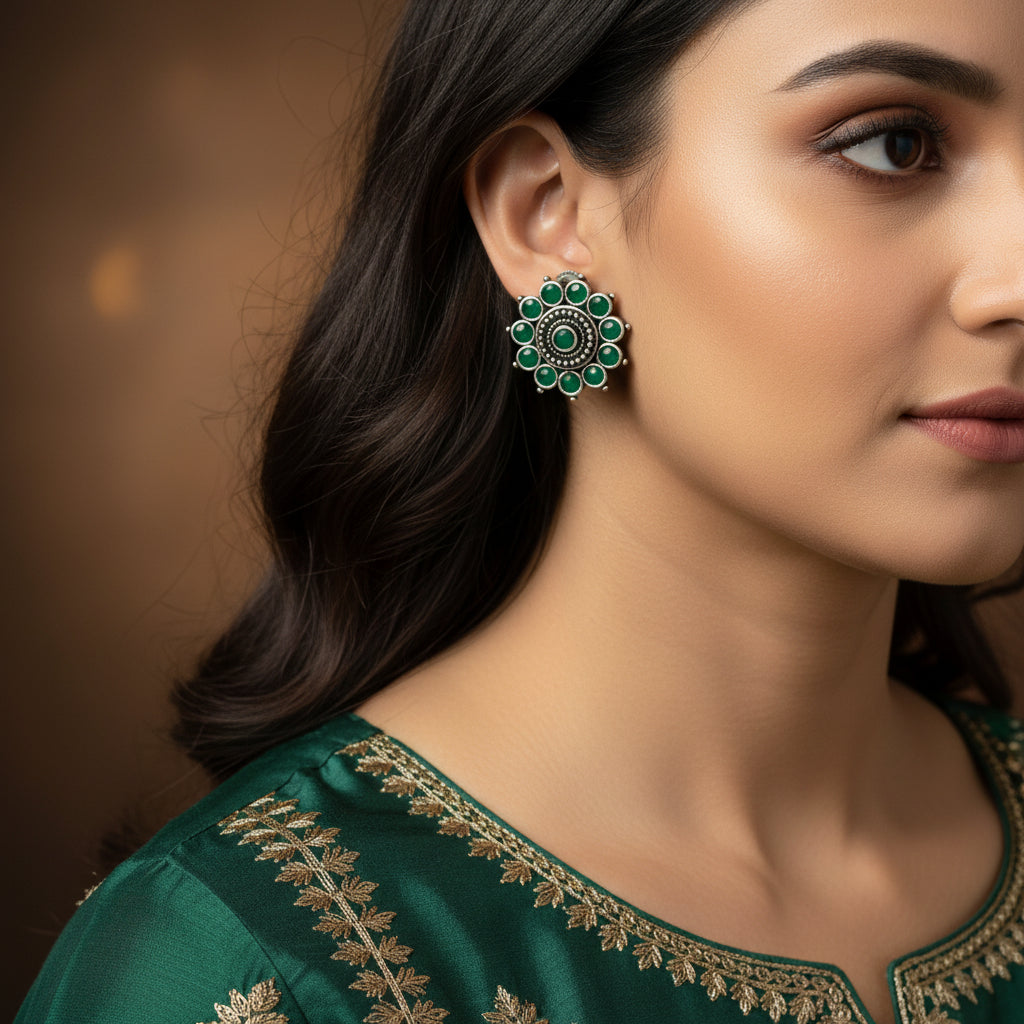 A pair of silver-toned circular stud earrings with green stone accents, displayed on a dark reflective surface.