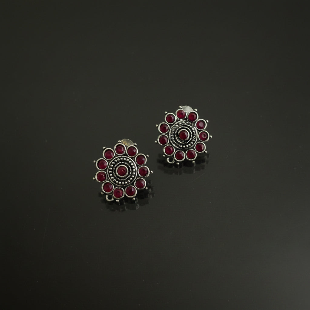 A pair of silver-toned Indian-style stud earrings with red stones, displayed on a dark reflective surface with the brand logo 'SAHELI BY BEST FRIEND' above them.