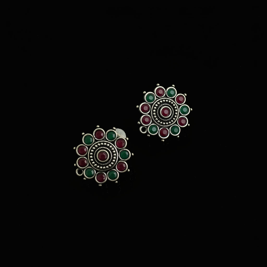 A pair of silver-toned Indian-style earrings with red and green stones, displayed on a dark reflective surface.