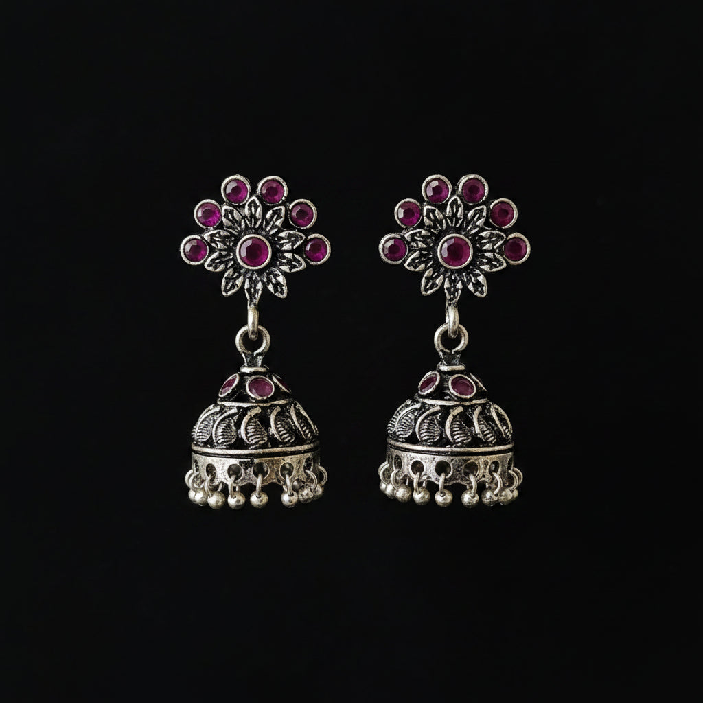 Silver jhumka earrings with red gemstones on a gray stand against a black background, branded 'Saheli'.