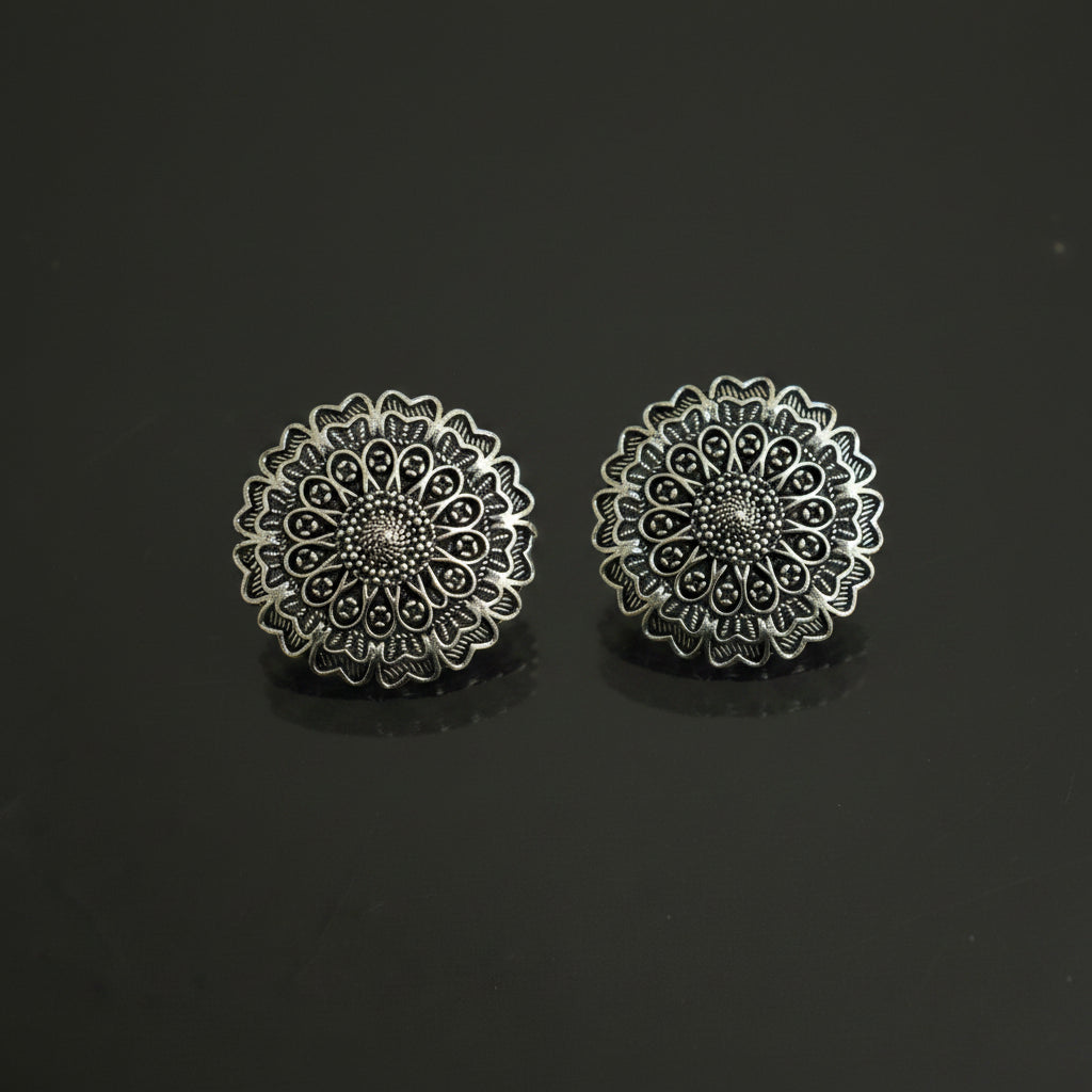A pair of round silver-toned filigree earrings with intricate openwork floral patterns, displayed on a dark reflective surface.