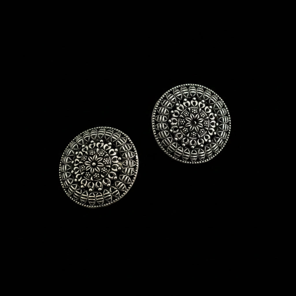 A pair of round silver-toned earrings with intricate floral and geometric filigree patterns, displayed on a dark reflective surface.