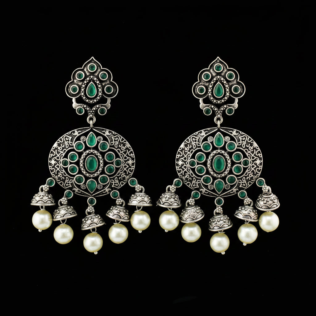 A pair of ornate silver-toned dangle earrings with green stone embellishments and white pearl drops, displayed on a gray stand against a gray background.