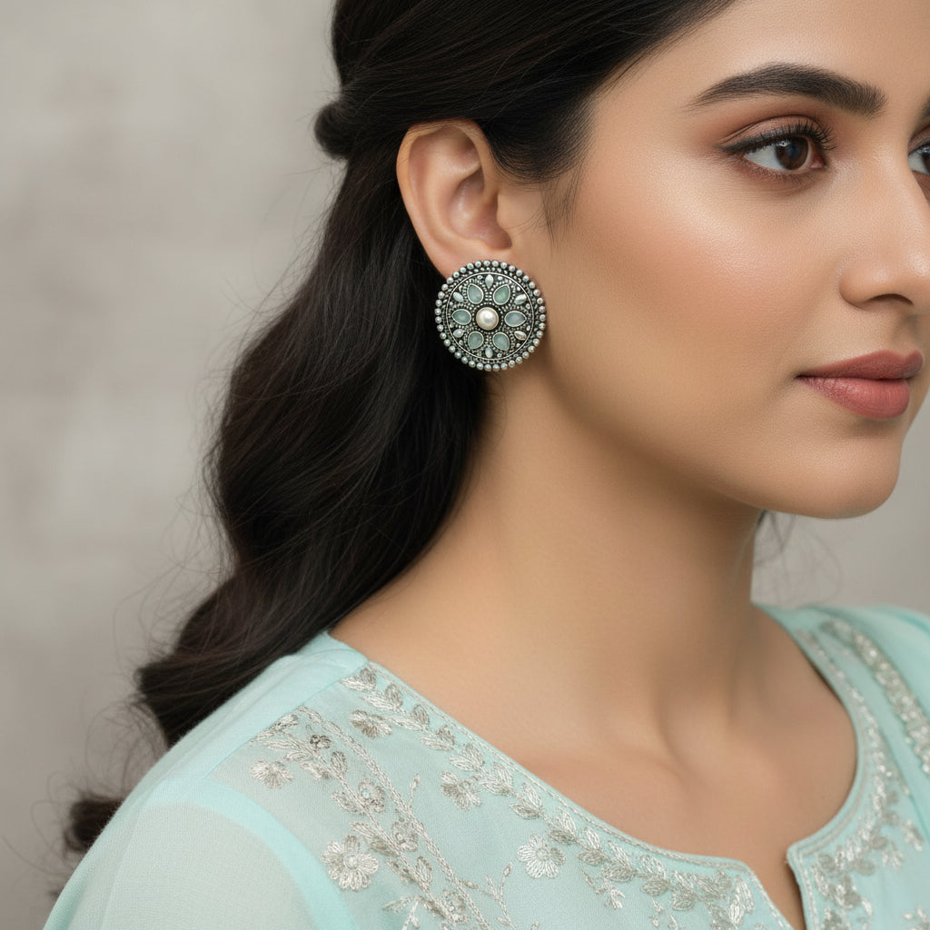 A pair of round silver-toned ornate brooches with turquoise-blue teardrop stones and a central pearl-like stone, displayed on a dark reflective surface.