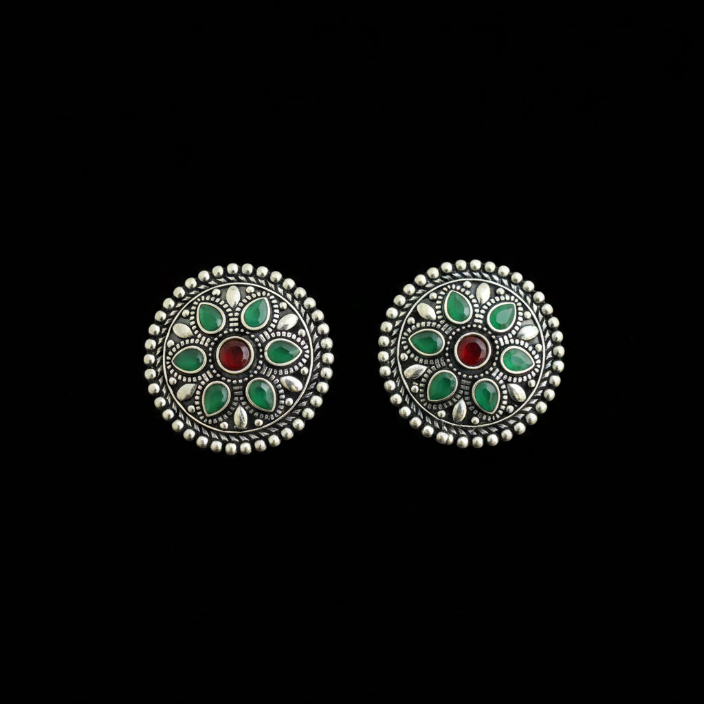A pair of round silver-toned earrings with intricate filigree detailing, featuring green stone inlays and a central red stone, displayed on a dark reflective surface.