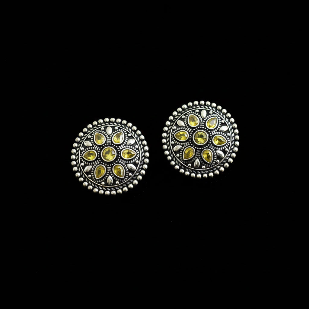 A pair of round silver-toned decorative buttons with intricate filigree detailing and small yellow gemstone inlays, displayed on a dark reflective surface.