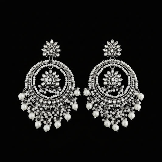 A pair of ornate silver-toned dangle earrings with intricate circular beadwork, floral motifs, and dangling pearl-like beads, displayed on a dark reflective surface.