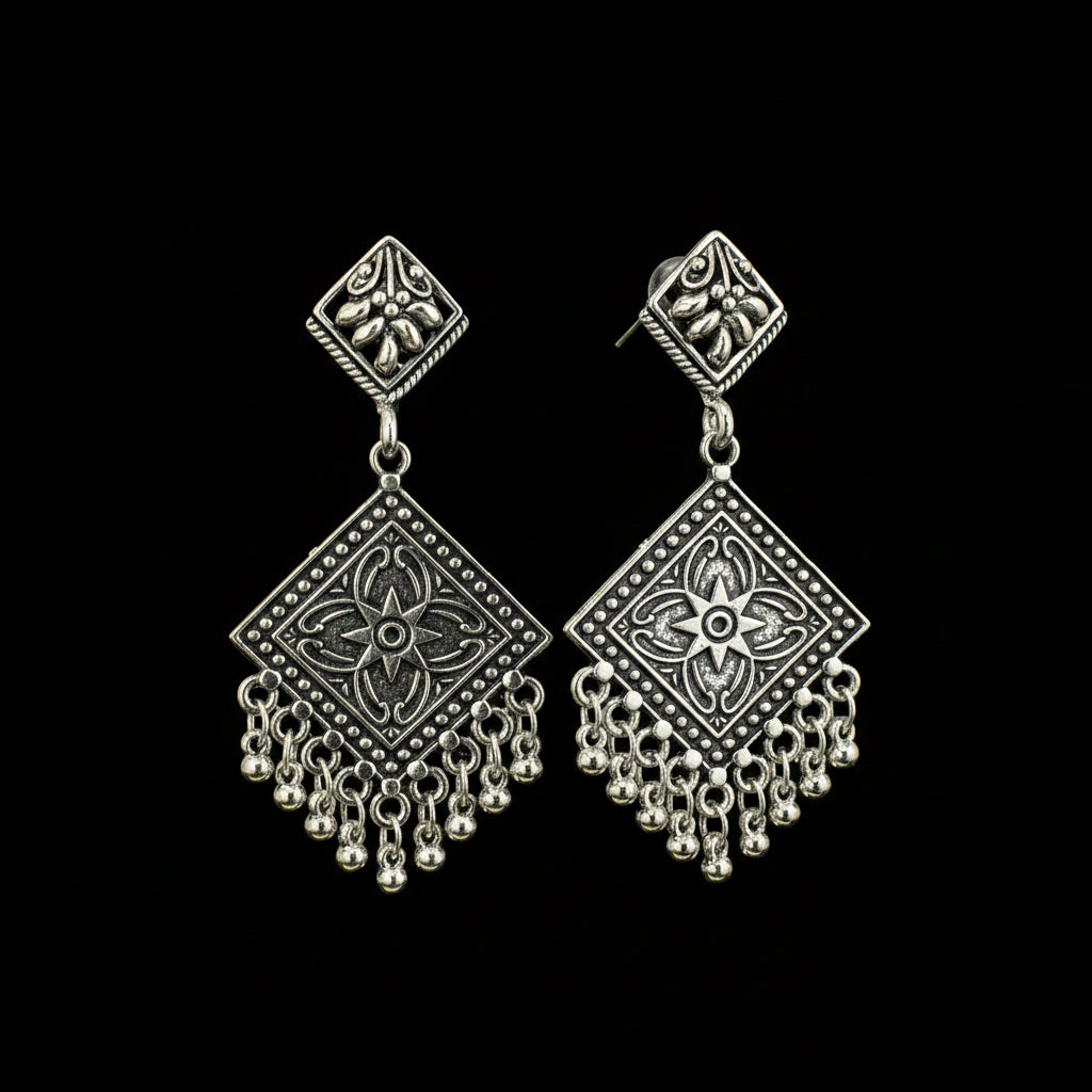 A pair of silver-toned geometric dangle earrings with ornate filigree and bead fringe, displayed on a black Y-shaped jewelry stand against a dark gray background.
