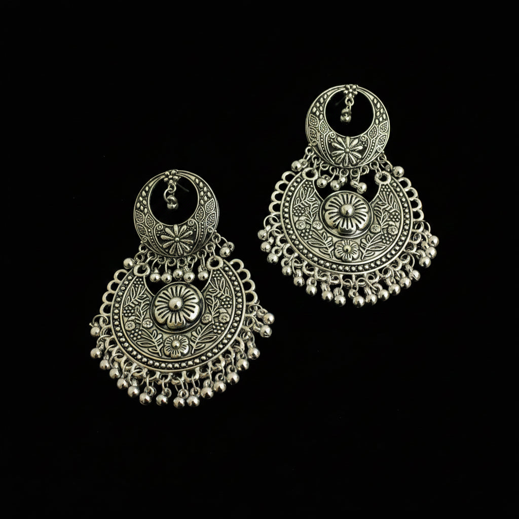 A pair of ornate silver-toned Indian jhumka earrings with intricate filigree work and dangling bead chains, displayed on a dark reflective surface.