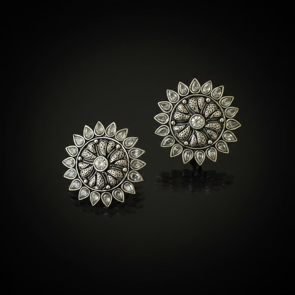 A pair of ornate silver-toned circular earrings with intricate filigree detailing and clear teardrop-shaped stones, displayed on a dark reflective surface.