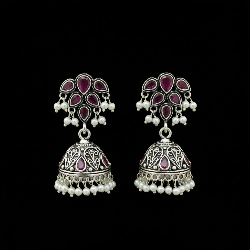 A pair of traditional Indian silver-toned dangle earrings with pink gemstone accents and white pearl bead borders, displayed on a black stand against a gray background.