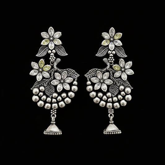 Silver floral earrings on a gray stand with 'Saheli' branding.
