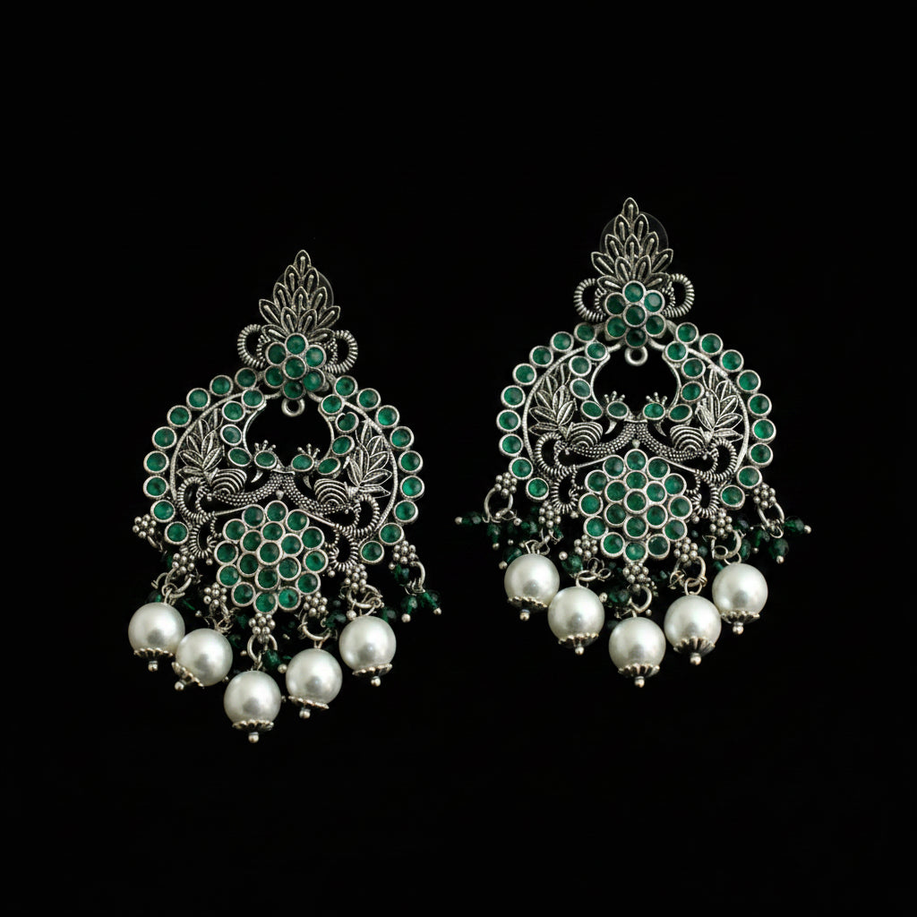 A pair of ornate silver-toned Indian jhumka earrings decorated with green stones and dangling white pearl beads, displayed on a dark reflective surface.