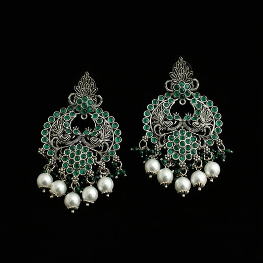 A pair of ornate silver-toned Indian jhumka earrings decorated with green stones and dangling white pearl beads, displayed on a dark reflective surface.