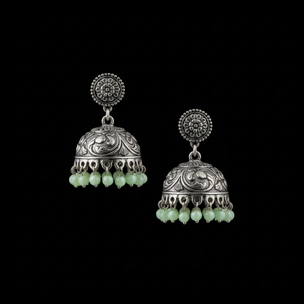 A pair of traditional Indian silver-toned jhumka earrings with green bead tassels, displayed on a gray velvet stand. The brand logo 'SAHELI MY BEST FRIEND' is visible at the top center.