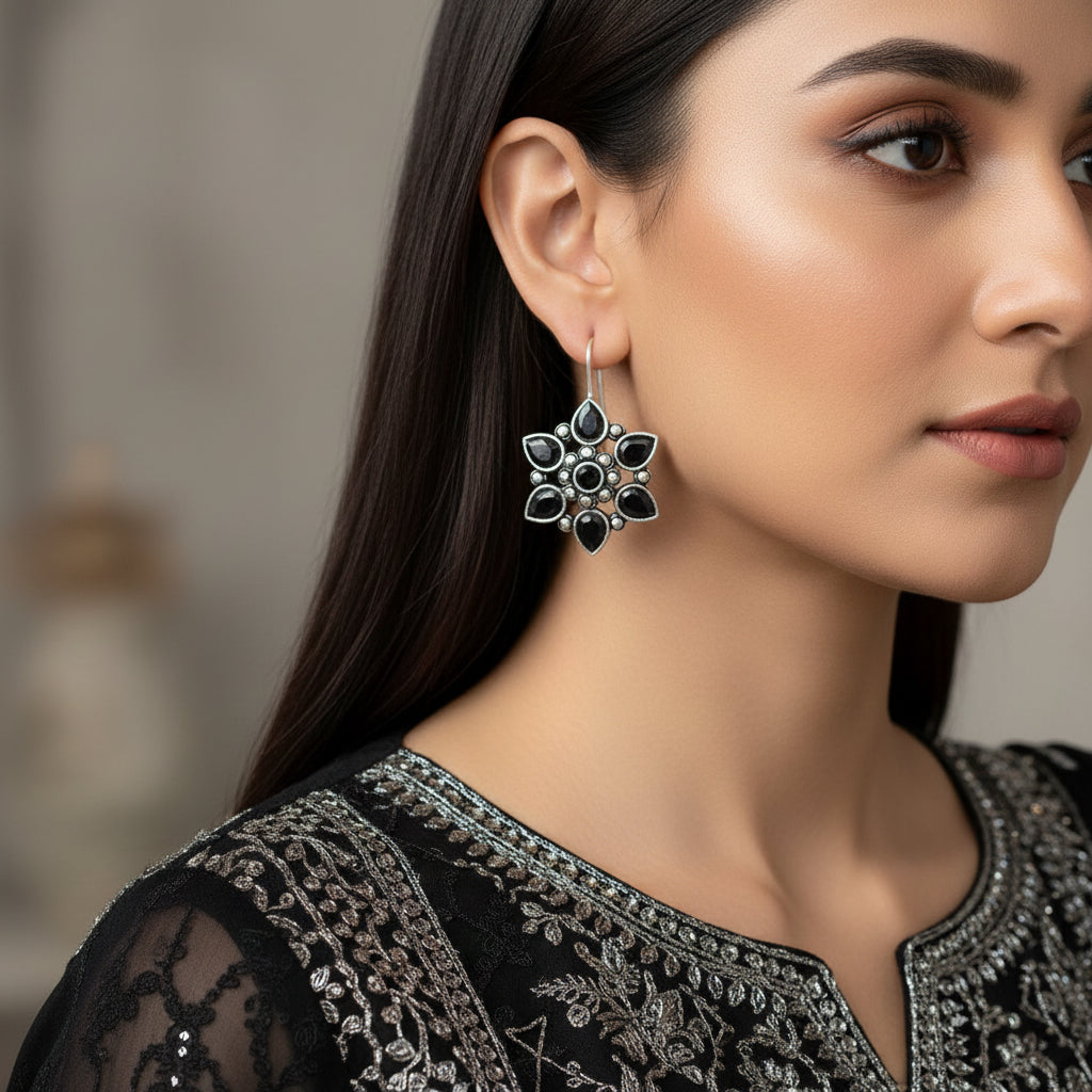 A pair of silver-toned flower-shaped drop earrings with dark teardrop gemstones and small silver dot accents, displayed on a gray cushion against a gray background.