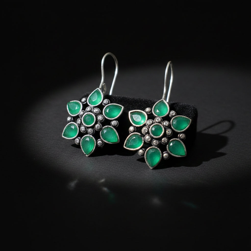 A pair of silver-toned dangle earrings with green teardrop-shaped stones and small round clear stones, displayed on a gray cushion against a dark gray background.