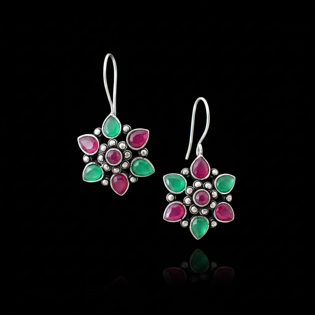 A pair of silver-toned flower-shaped earrings with green and pink teardrop-shaped stones, displayed on a gray cushion against a dark gray background.