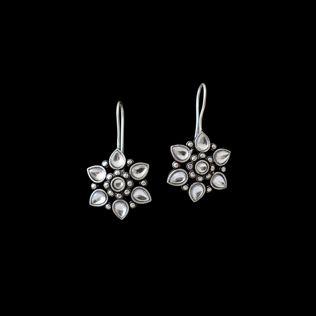 A pair of silver-toned dangle earrings with floral starburst designs, featuring clear teardrop-shaped stones and small silver beads, displayed on a gray velvet jewelry stand against a dark gray background.