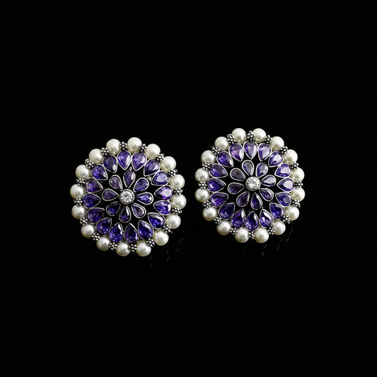 A pair of round floral stud earrings featuring purple amethyst teardrop stones, a central clear crystal, and a border of white faux pearls on silver-toned settings, displayed on a dark reflective surface with the SAHELI brand logo above.