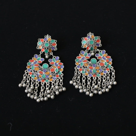 A pair of ornate silver-toned Indian chandbali earrings decorated with multicolored enamel in green, orange, red, and purple, featuring dangling silver chains and bead clusters on a dark reflective surface.